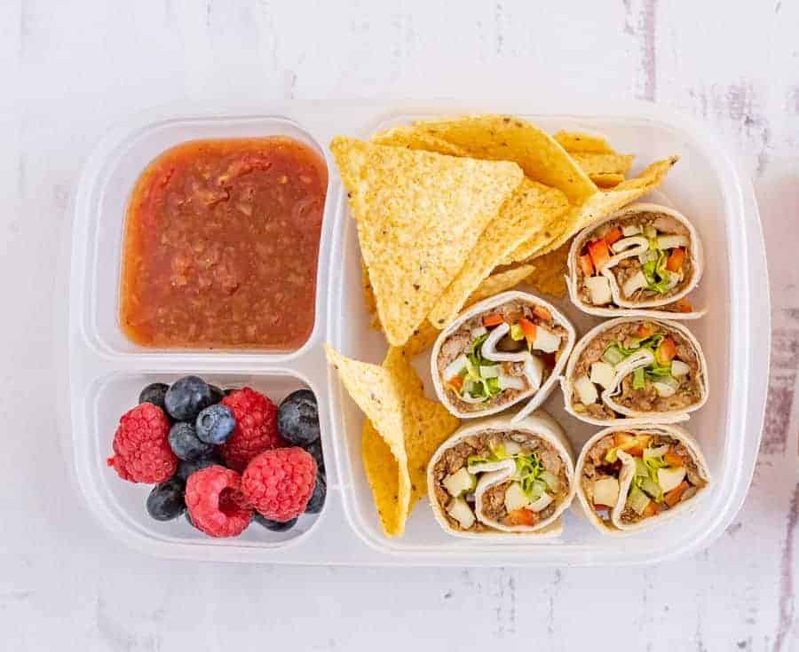 A lunchbox with easy pinwheel recipes—tortilla wrap pinwheels filled with veggies—plus tortilla chips, salsa, and a side of fresh raspberries and blueberries.
