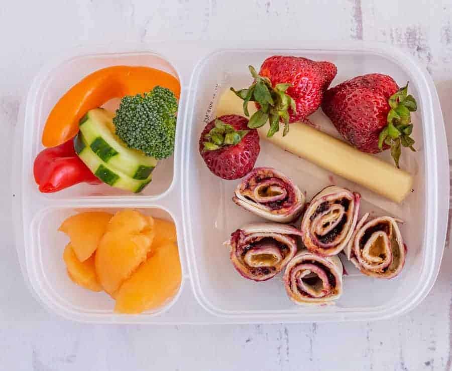 A divided lunch container with strawberries, string cheese, easy pinwheel recipes made as tortilla roll-ups with a spread, sliced cucumber, broccoli, mini sweet peppers, and cantaloupe chunks on a white surface.