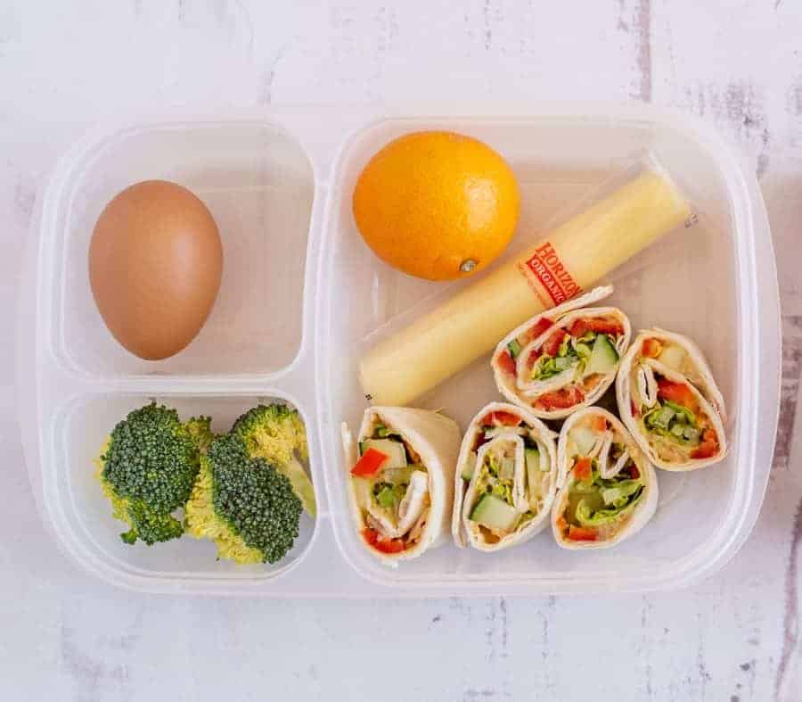 A lunchbox with sections holding a hard-boiled egg, broccoli florets, an orange, a cheese stick, and turkey and veggie pinwheel wraps—perfect for enjoying easy pinwheel recipes on the go.