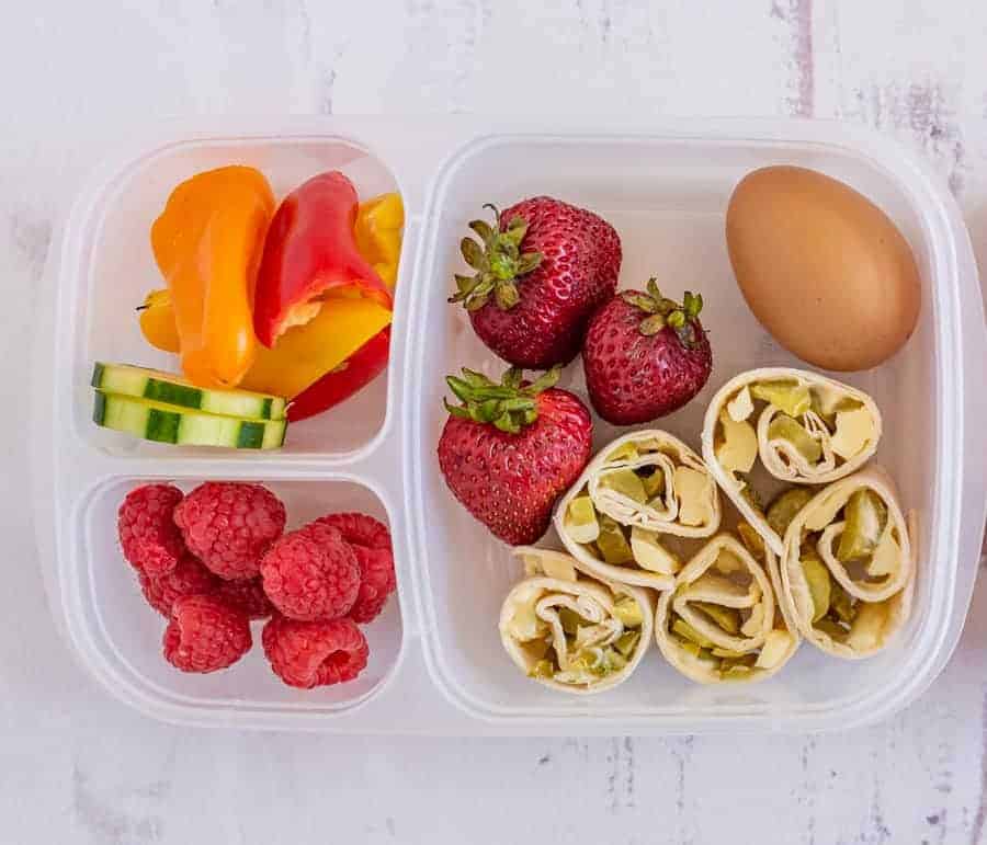 A lunchbox with sliced mini bell peppers, cucumber, strawberries, a hard-boiled egg, fresh raspberries, and rolled tortilla pinwheels with a veggie filling—perfect for those seeking easy pinwheel recipes for quick meals.