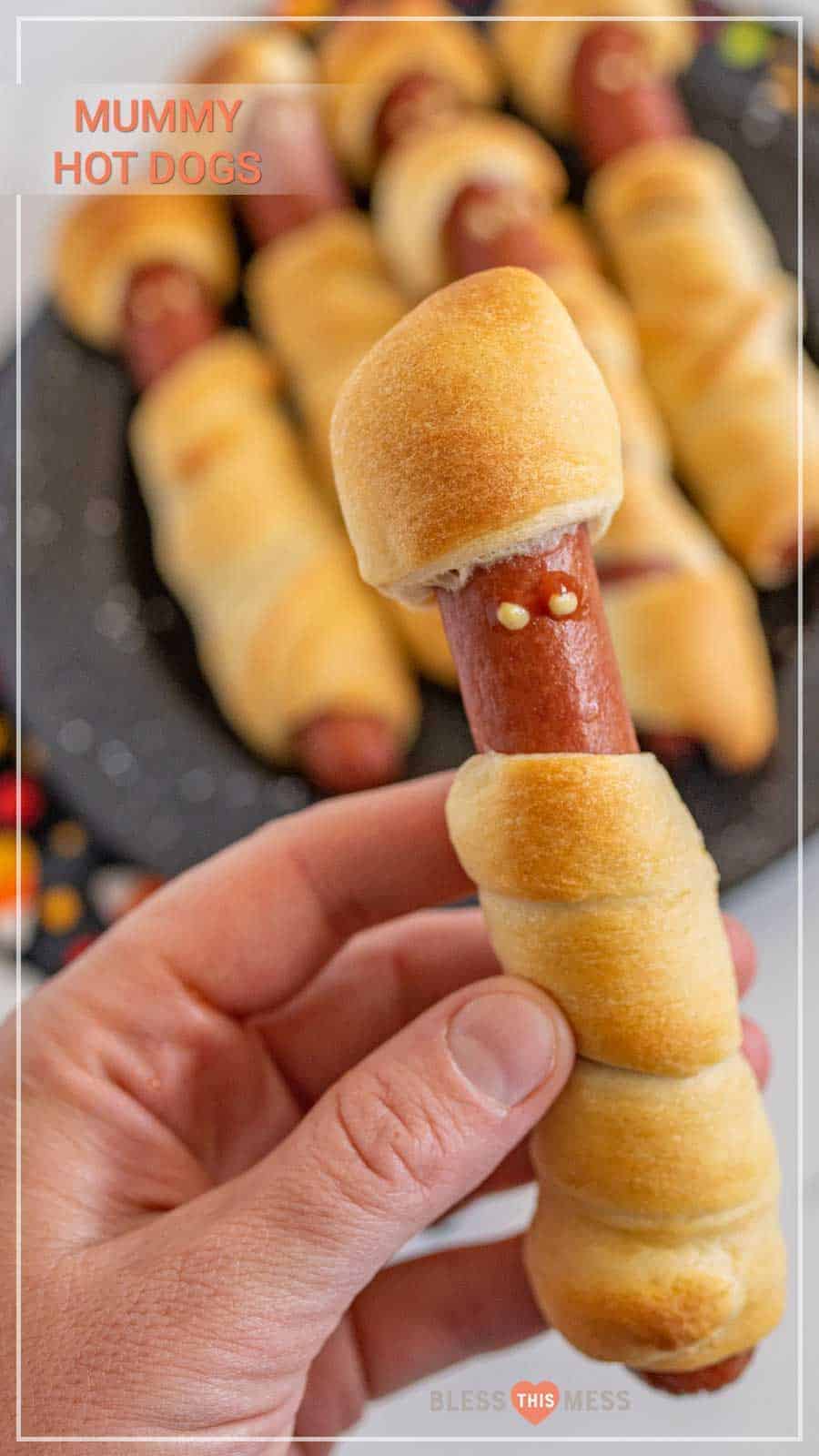 hand holding a hot dog wrapped in a crescent roll to look like a mummy more mummy hot dogs blurred in background.