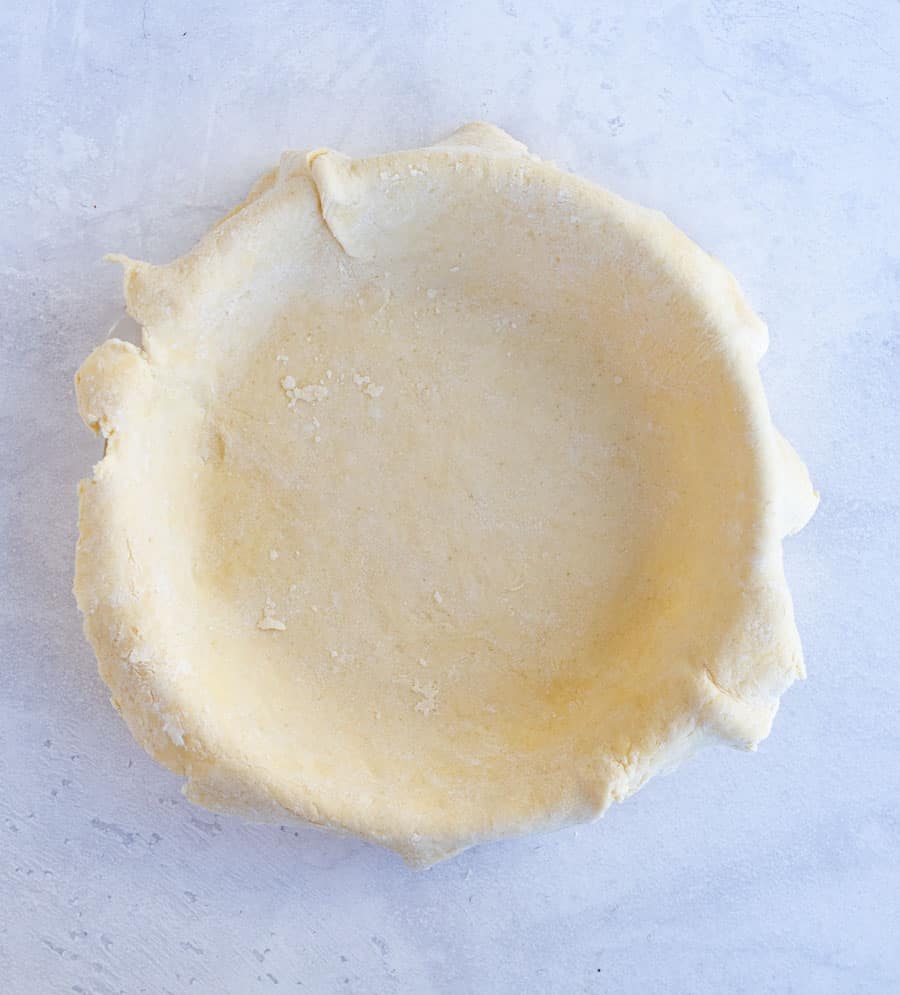 An all butter pie crust is neatly placed in a round pie dish on a light gray surface. The edges of the dough are slightly overhanging the dish, ready for trimming or crimping.
