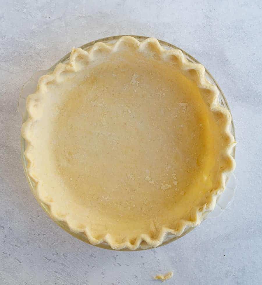 A clear glass pie dish with uncooked all-butter pie crust, featuring crimped edges, sits on a light, textured surface. The buttery crust is evenly spread and gently pressed into the dish, ready for filling or baking.