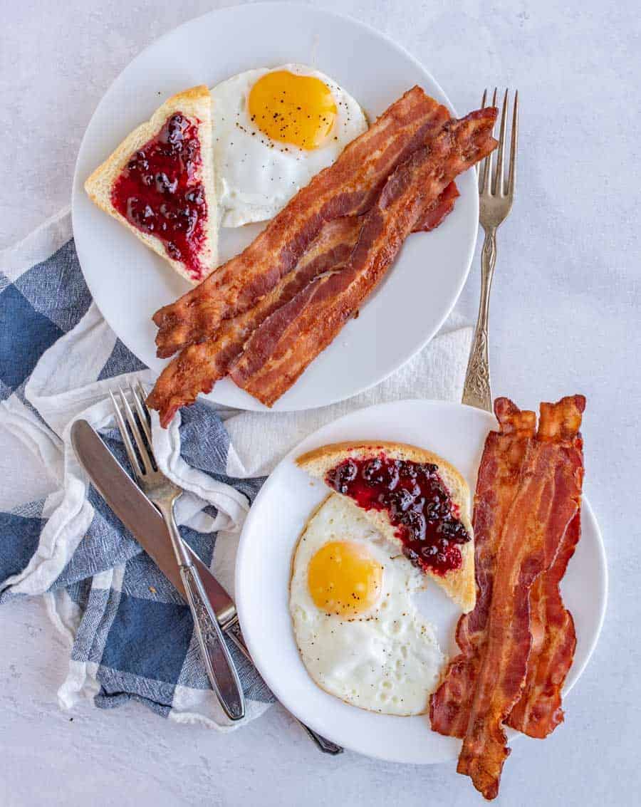 Top view of two white plates each with three slices of bacon, a fried egg, and toast with jam on it.