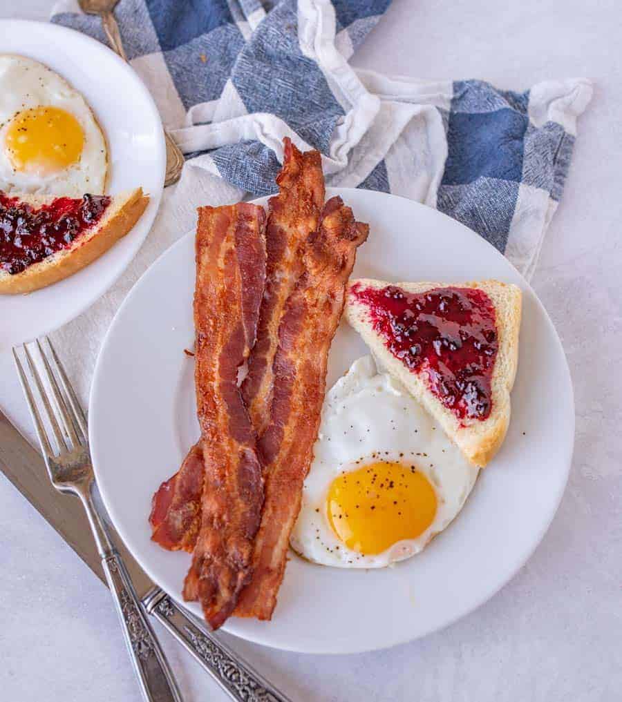 Up close of a plate with three pieces of bacon, an egg sunny side up, and a piece of toast with jam on it.