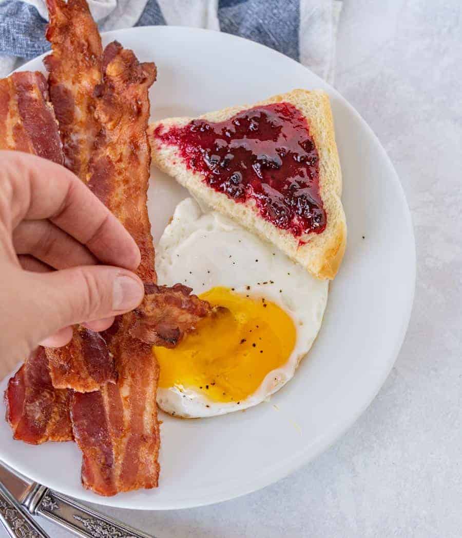 A hand dipping a piece of bacon in the runny egg yolk next to toast with jam.