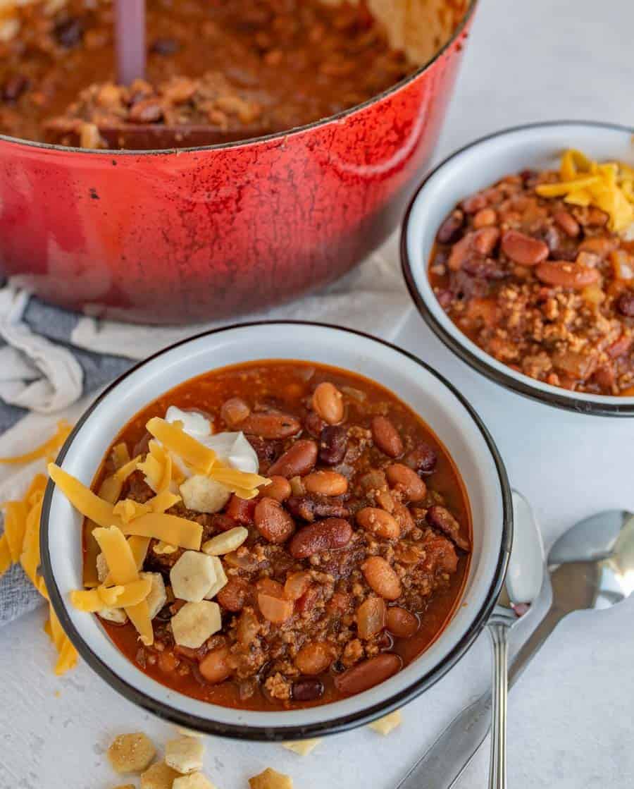 two bowls of chili in white bowls with cheese and oyster crackers on top red dutch oven full of chili in background.