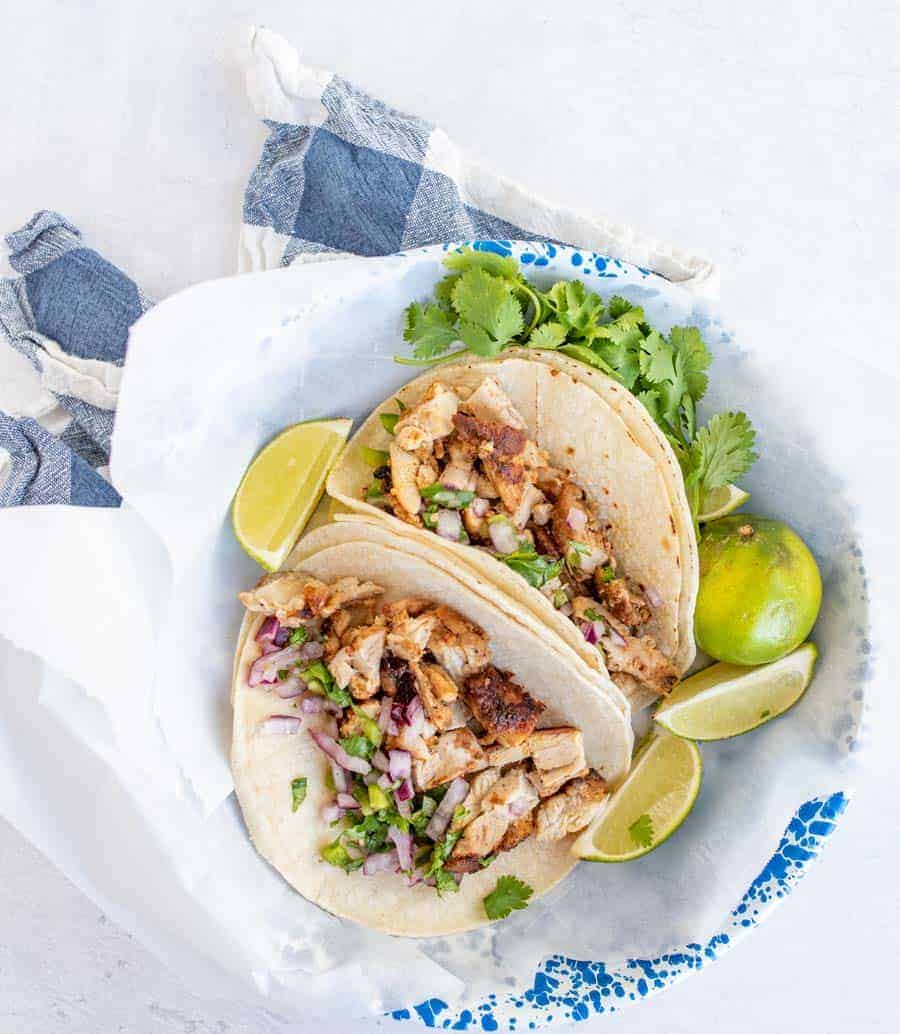 two chicken tacos, some lime wedges, and cilantro on a plate.
