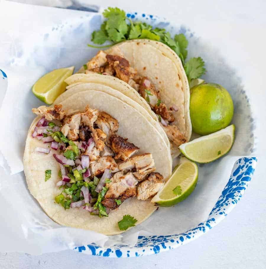 chicken tacos in two tortillas in bowl with cilantro and lime wedges.
