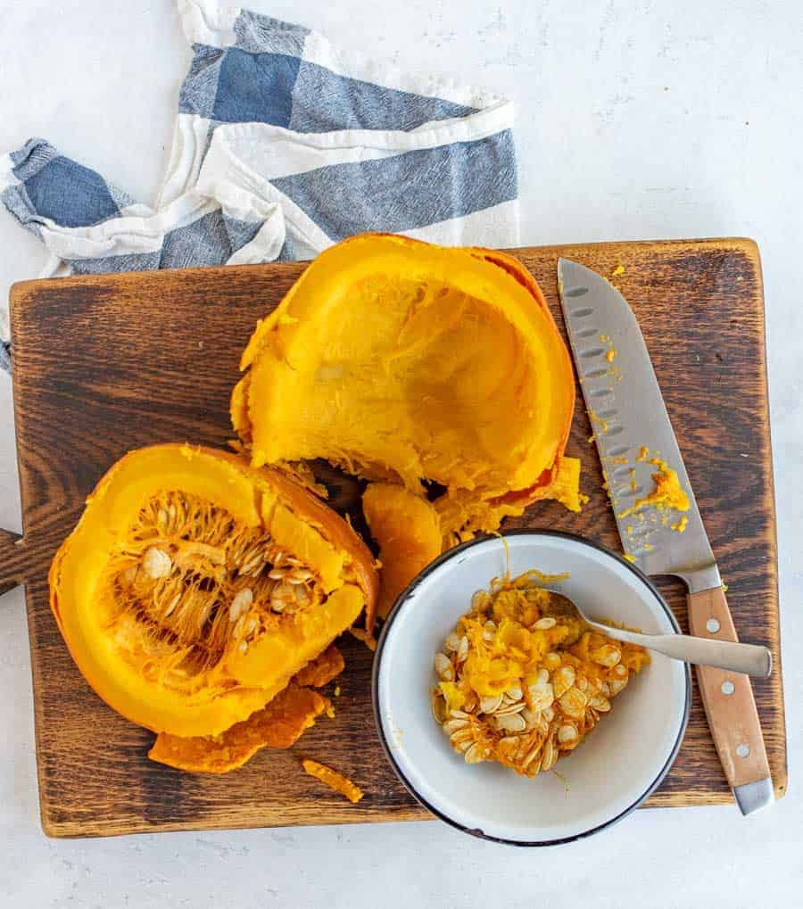wooden cutting board with cut open orange pumpkin puree and seeds in a white bowl butcher knife on cutting board.
