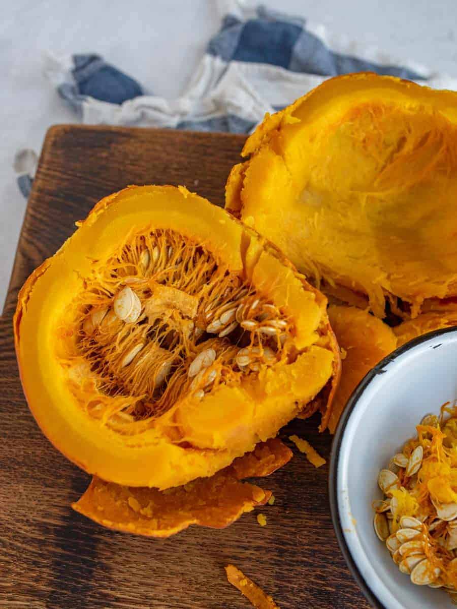 wooden cutting board with cut open orange pumpkin puree and seeds in a white bowl on cutting board.
