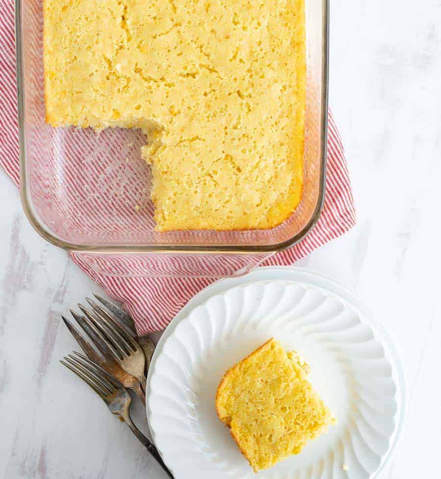 top view of corn casserole in a square glass dish beside white place with a piece of corn casserole on it.
