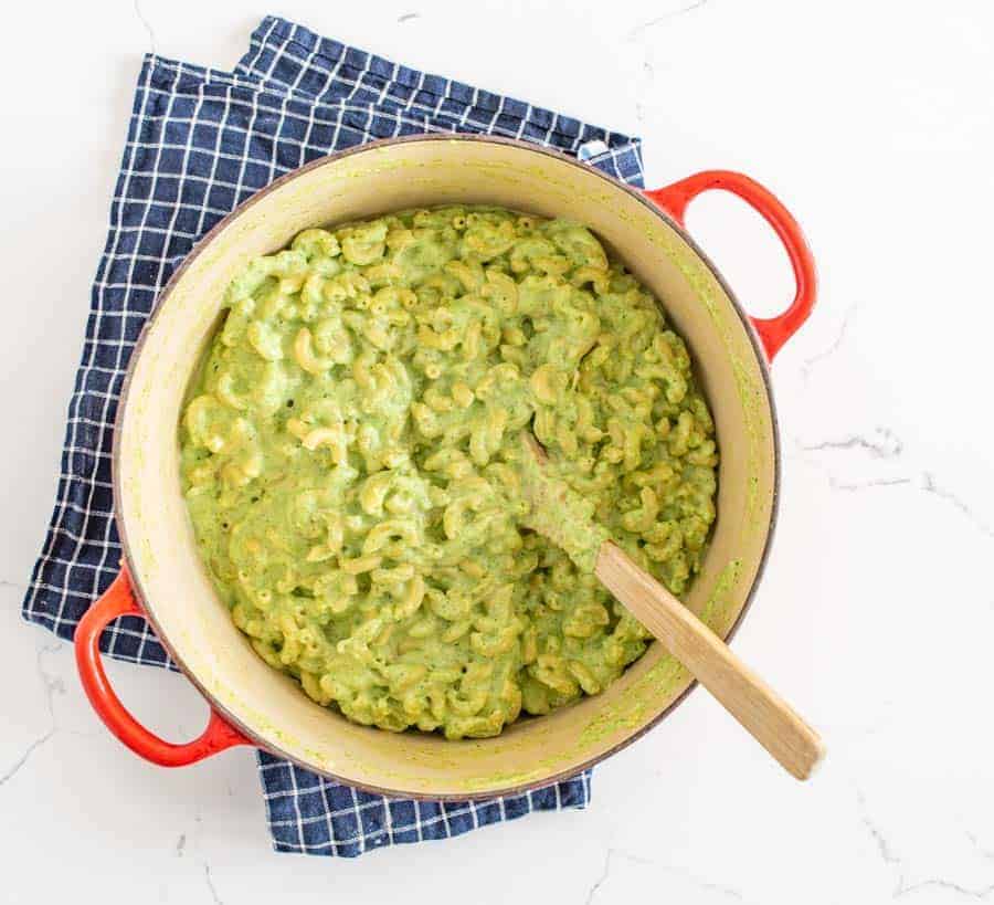 green macaroni and cheese in a red dutch oven pot with a wooden spoon stuck in the pot sitting on blue and white towel on a white marble counter.
