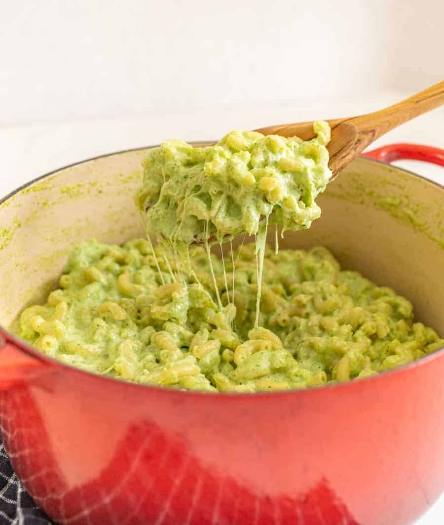 green mac and cheese in a red dutch oven wooden spoon holding up a gooey bite.