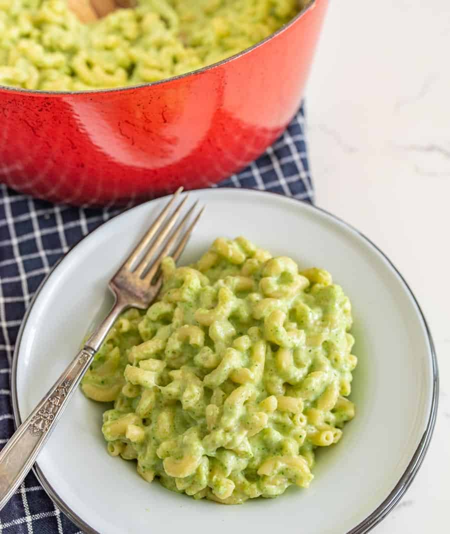 green macaroni and cheese in a white bowl with a metal fork red dutch oven pot full of mac and cheese in the background.