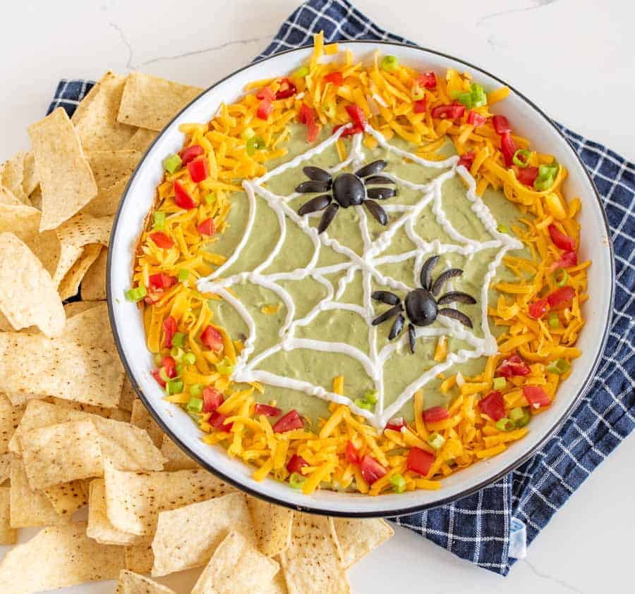 bean dip with green guacamole in a white dish edges covered in cheese tomatoes and peppers with a sourcream spider web piped on and two spiders made out of olives tortilla chips on the side.