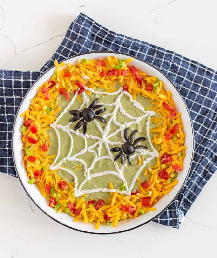 bean dip with green guacamole in a white dish edges covered in cheese tomatoes and peppers with a sourcream spider web piped on and two spiders made out of olives.