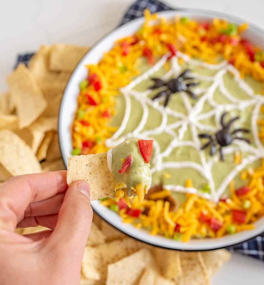 bean dip with green guacamole in a white dish edges covered in cheese tomatoes and peppers with a sour cream spider web piped on and two spiders made out of olives a hand holding a chip with dip.