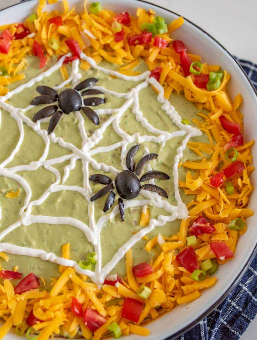 Halloween bean dip with green guacamole in a white dish edges covered in cheese tomatoes and peppers with a sourcream spider web piped on and two spiders made out of olives.