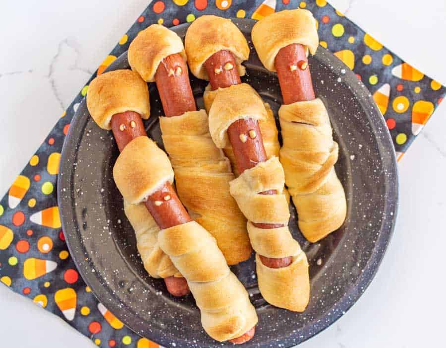 a plate of crescent dough-wrapped hot dogs with mummy faces with a candy corn napkin underneath.