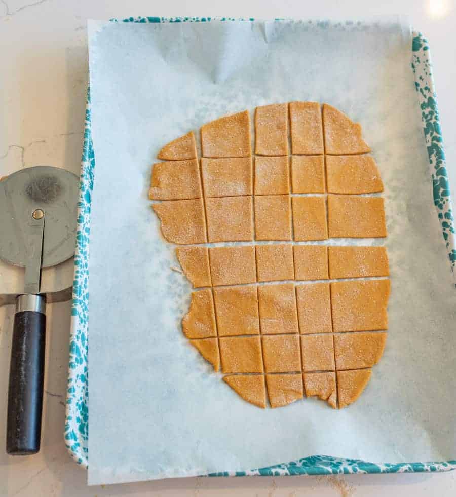 A baking sheet lined with parchment paper holds rolled-out pumpkin dog treats dough cut into squares. A pizza cutter with a black handle lies to the left of the tray on a white countertop.
