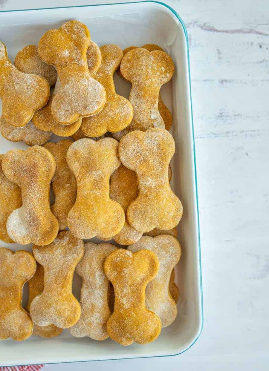 A tray filled with bone-shaped, golden-brown pumpkin dog treats, lightly dusted with flour and arranged neatly in overlapping rows.