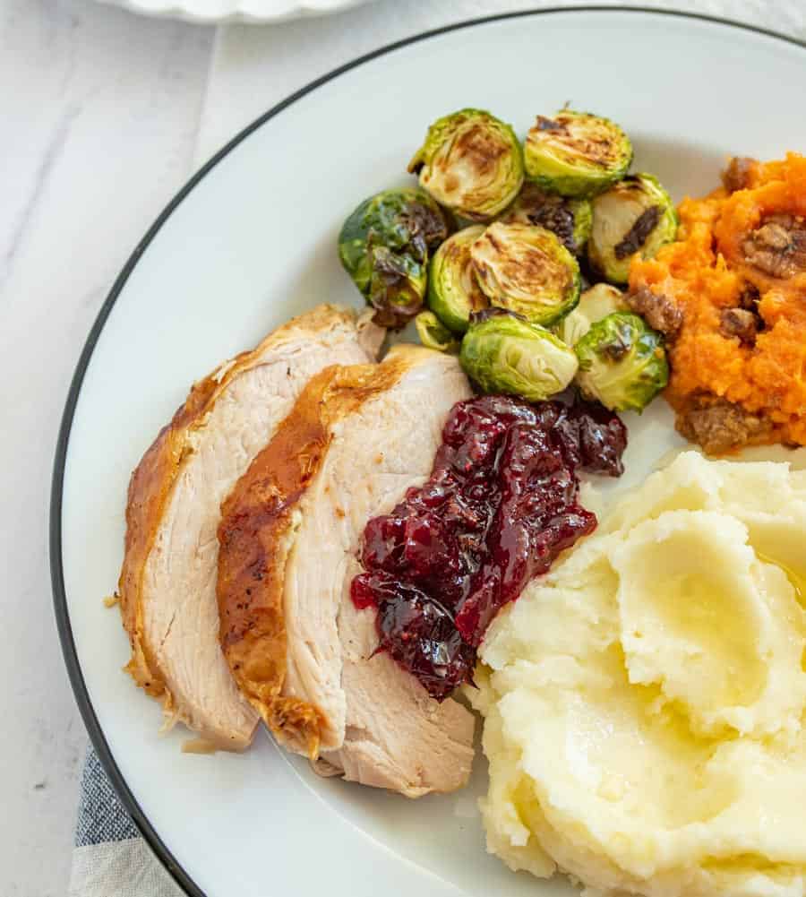 Roasted turkey slices, roasted brussel sprouts, cranberry sauce, mashed potatoes, and sweet potato casserole all on a white plate.
