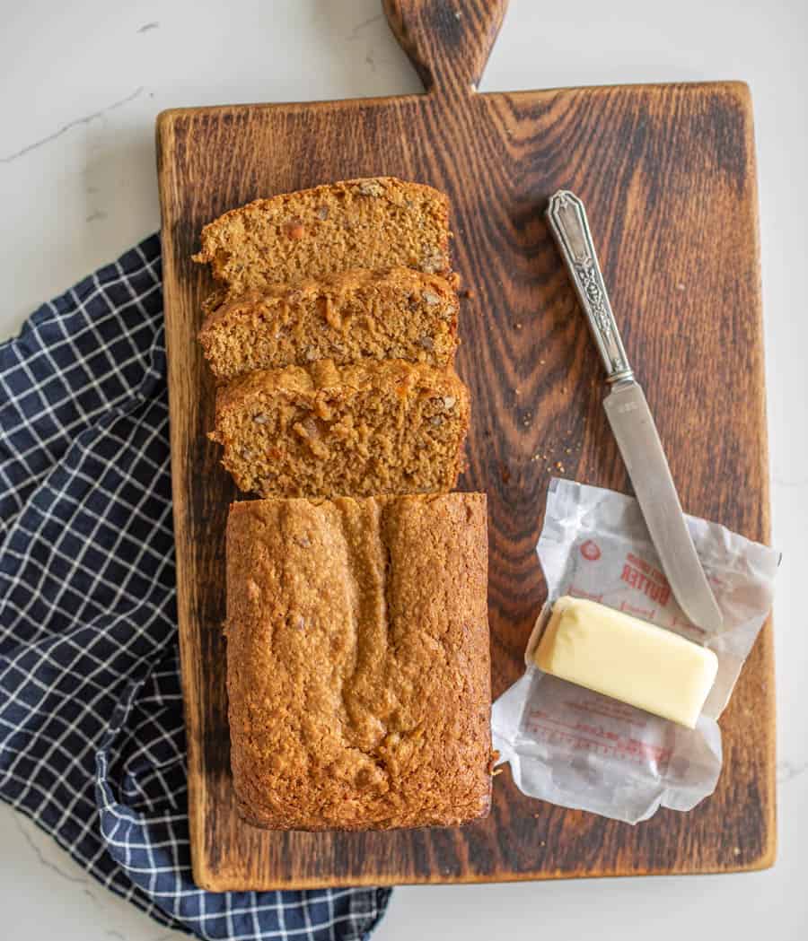a loaf of sweet bread sitting wooden cutting board on a navy blue and white checkered dish towel half a stick of butter and a butter knife beside the bread.