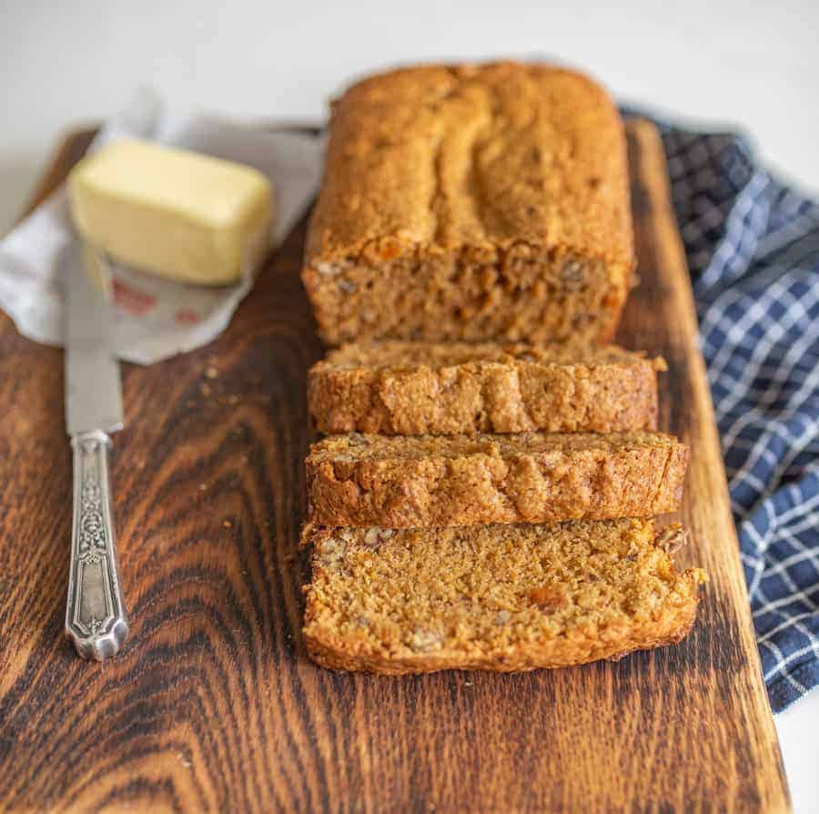 a loaf of sweet bread sitting wooden cutting board on a navy blue and white checkered dish towel half a stick of butter beside the bread.