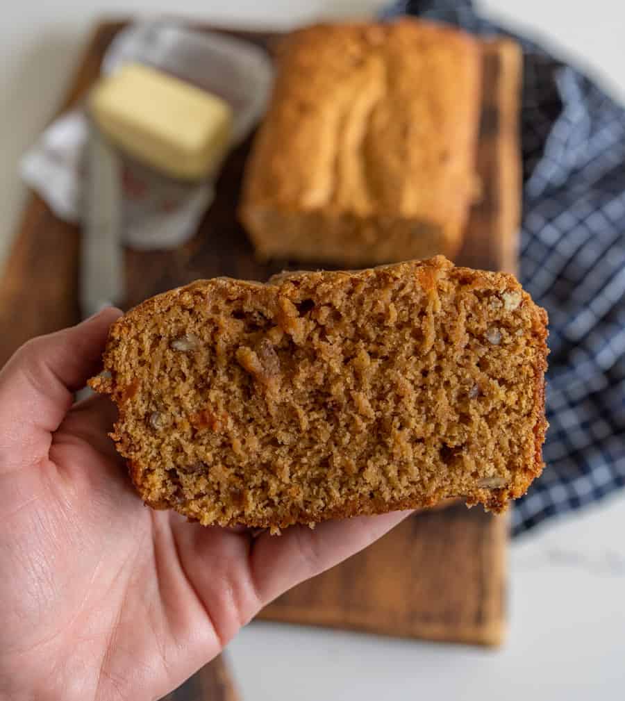 a hand holding a slice of sweet potato bread with pecans butter and butterknife in background.