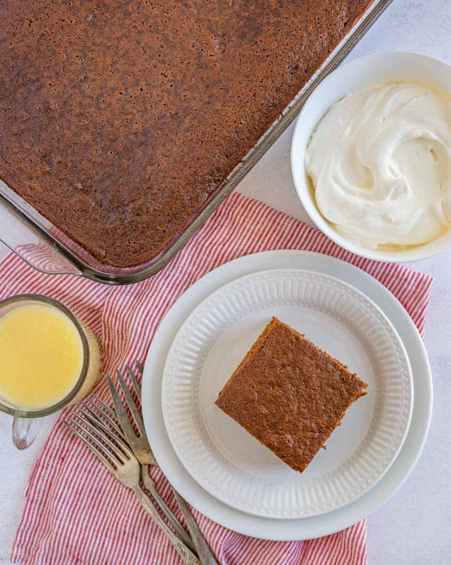 A pan of plain brown cake, a bowl of melted butter, 3 forks, a white plate with a slice of plain brown cake, and a bowl of whipped cream. All on a white background with a red and white striped towel.