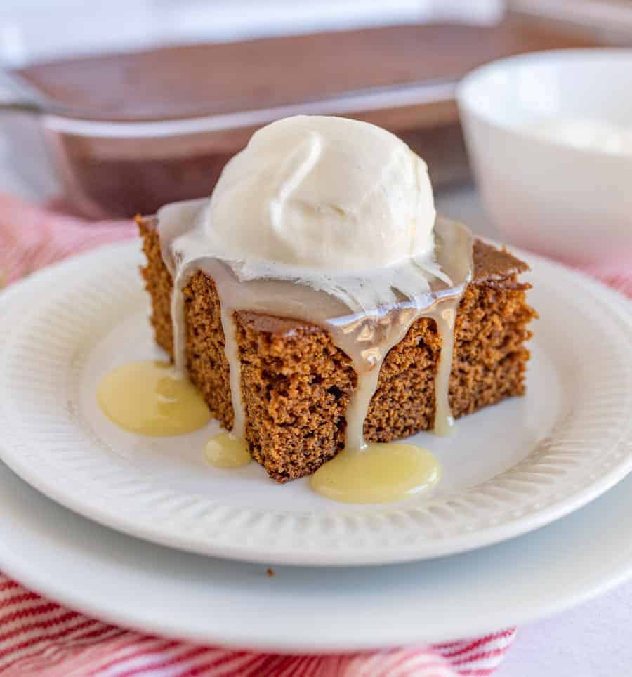 A slice of gingerbread cake served on a white plate. The cake is topped with caramel sauce and a scoop of vanilla icecream.