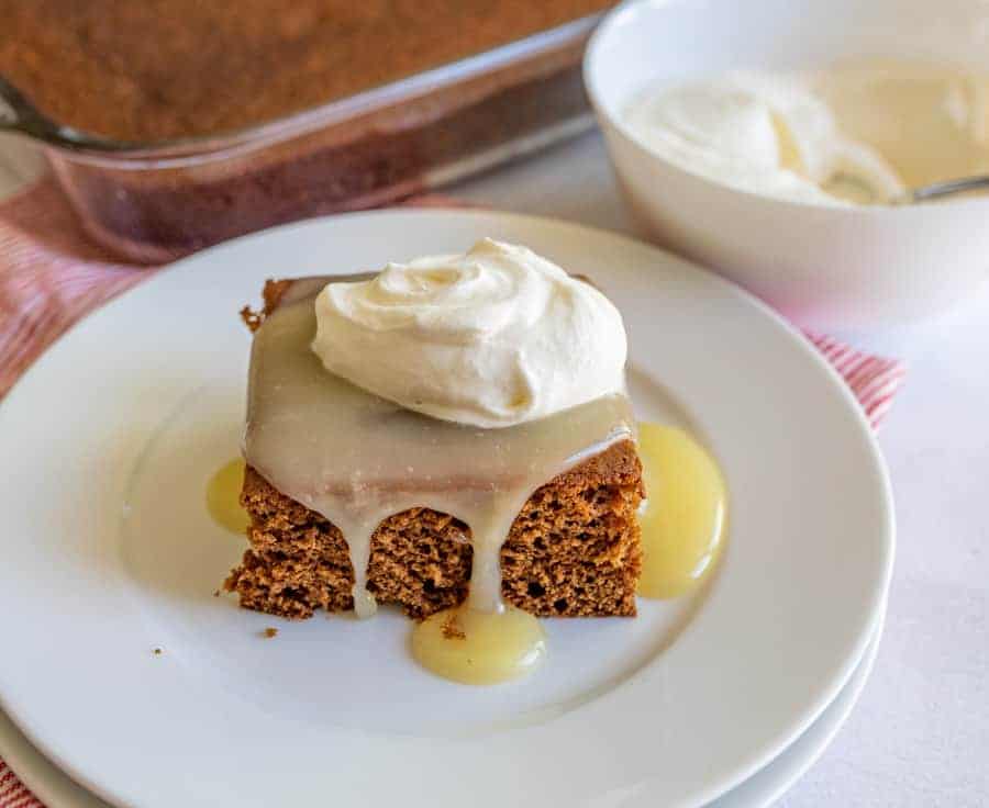 A slice of gingerbread cake with caramel and whipped cream on top, placed on a white plate.