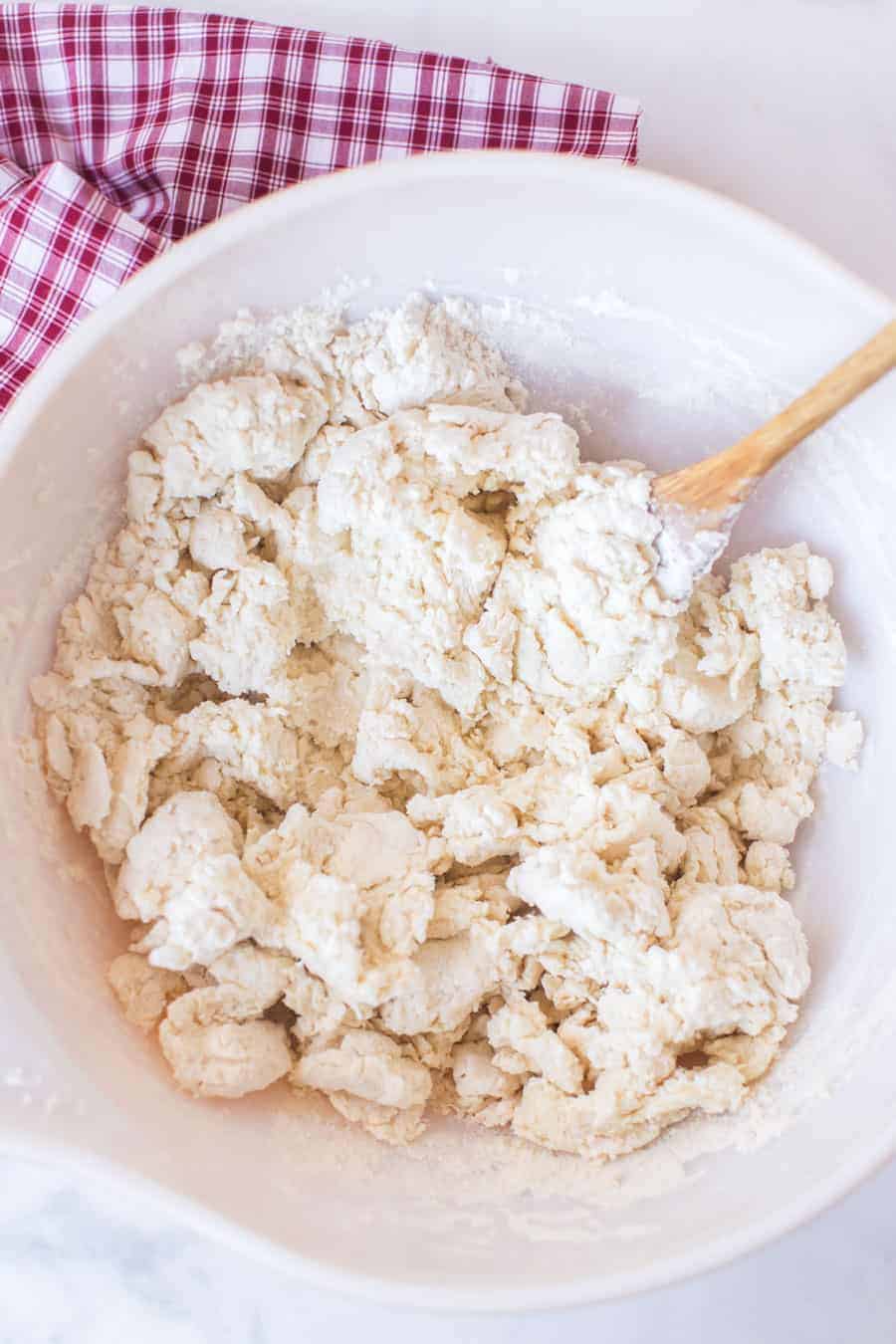 biscuit dough in white mixing bowl with wooden spoon on red and white checked towel.