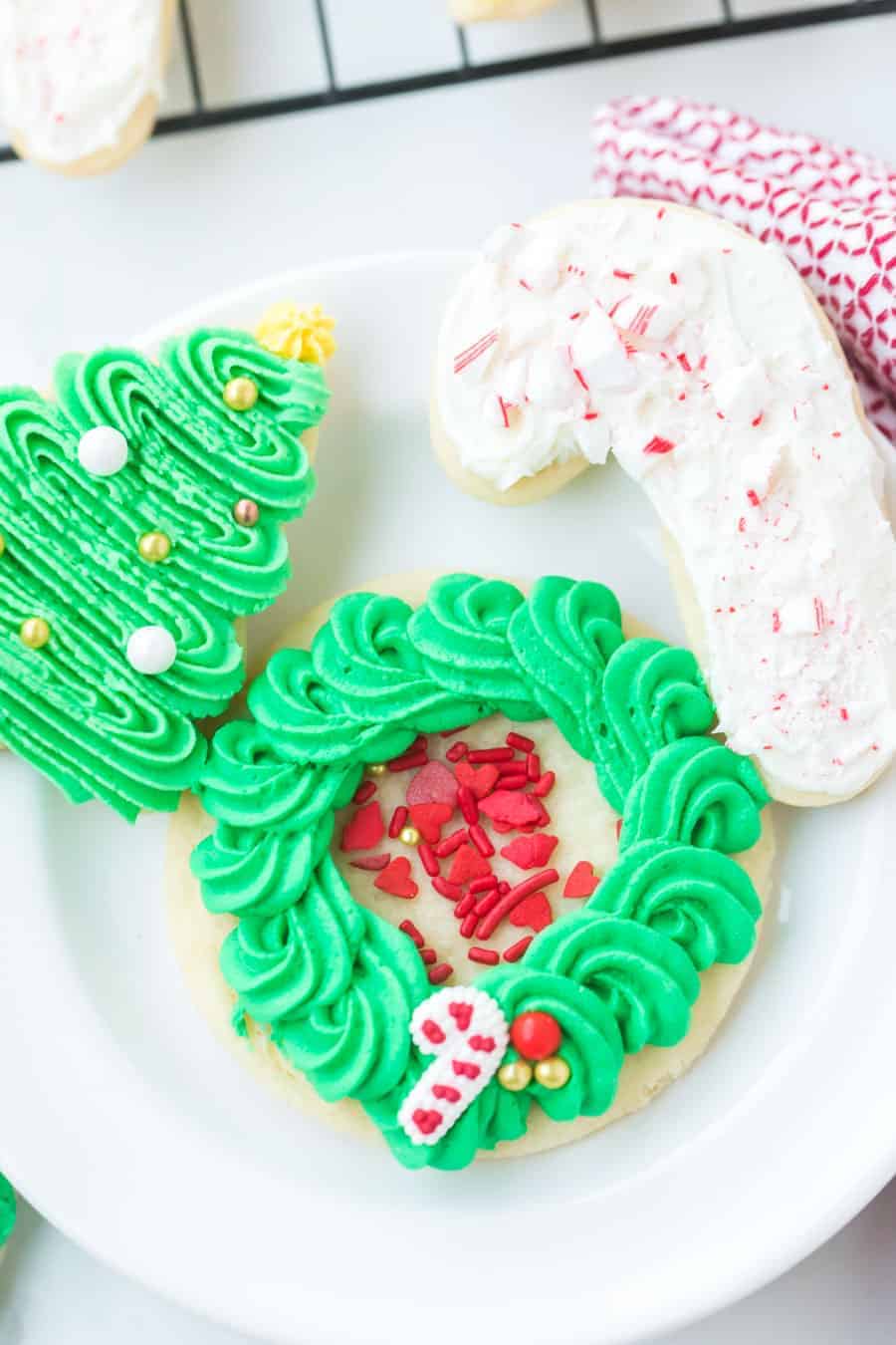 Iced sugar cookies in the shape of trees, wreaths, and candy canes on a white plate.
