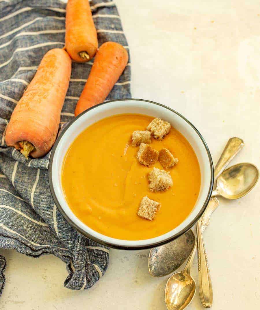 photo of a bowl of creamy carrot soup with a row of croutons in it a raw whole carrots in the background.
