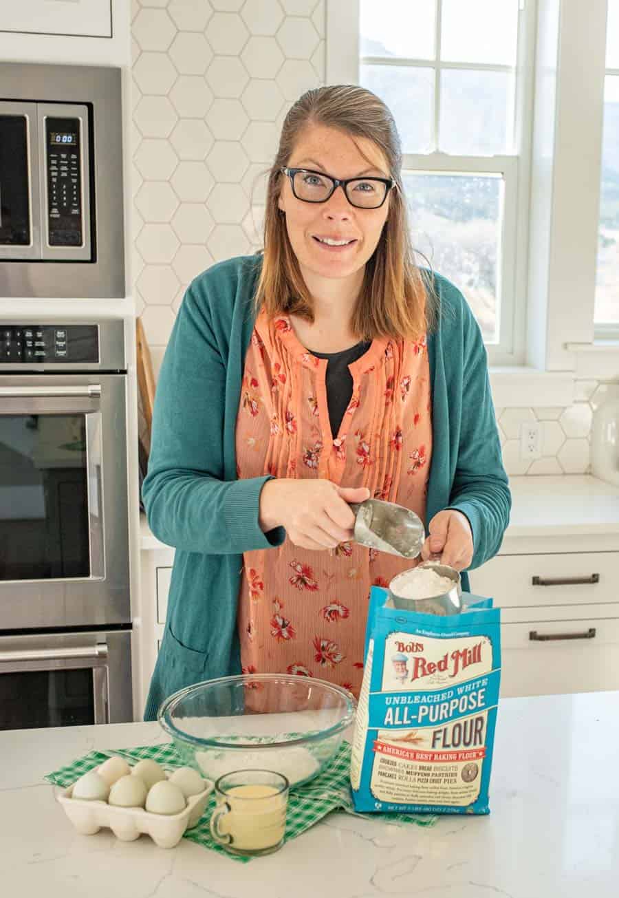 A woman in a white kitchen scooping flour out of the bag and placing it in a glass bowl. A carton of eggs and a glass of melted butter are on the island with a green towel.