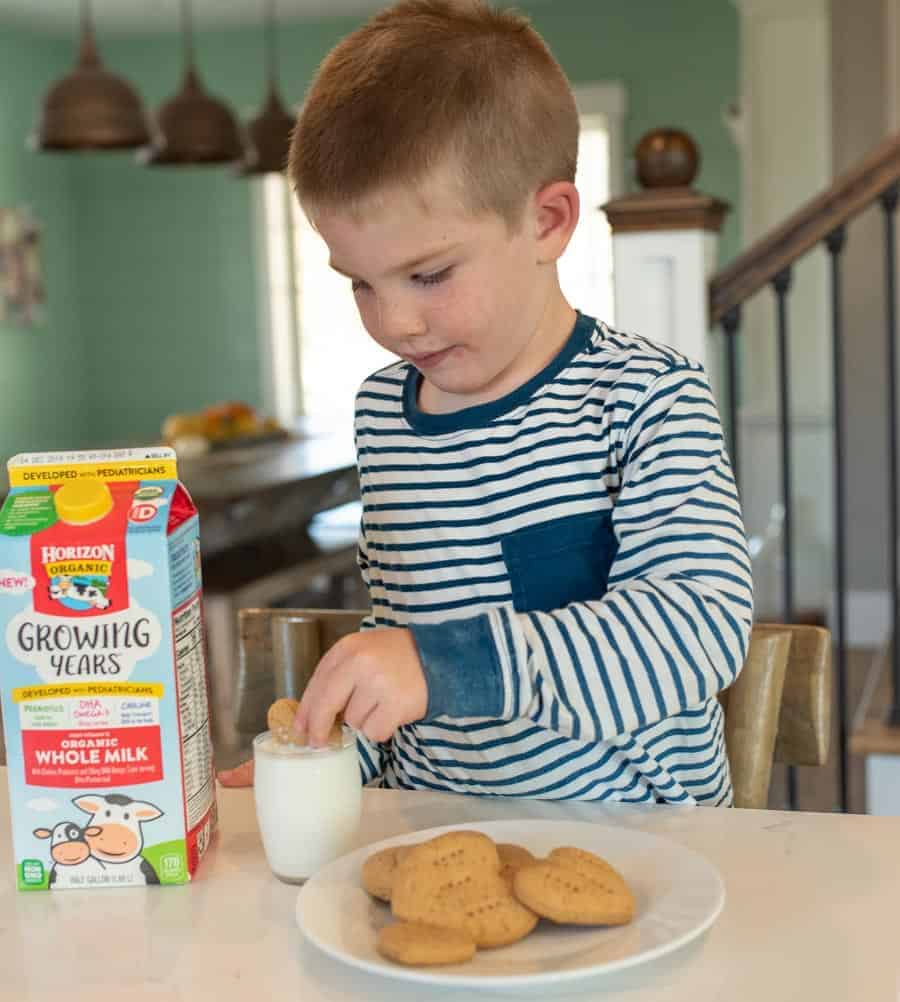 little boy with brown hair in a blue and white striped shirt dipping a heart-shaped graham cracker into a glass of milk.