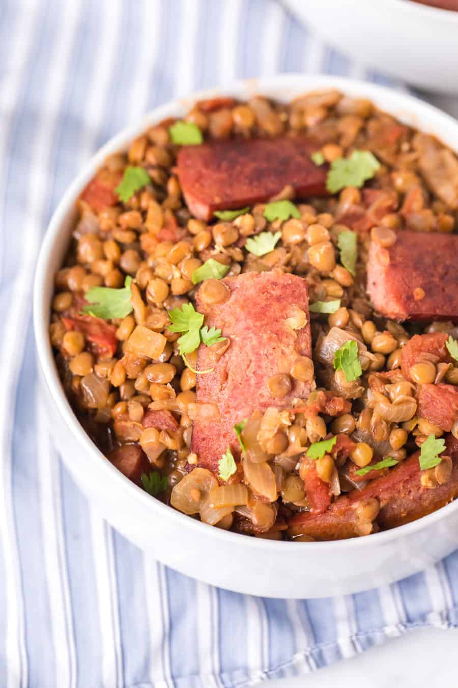 closeup image of spicy lentils and sausage in white bowl on blue and white striped towel.