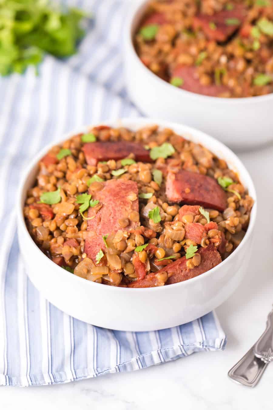 2 white bowls of spicy lentils and sausage with a spoon.