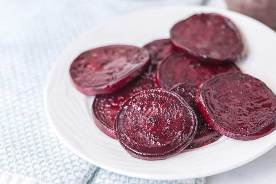 A white plate holds several slices of roasted beets, arranged in overlapping layers. The deep purple, slightly glossy beets contrast beautifully against the light, textured background.