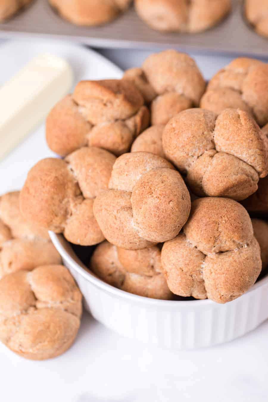 whole wheat clover rolls in white dish next to stick of butter on white surface.