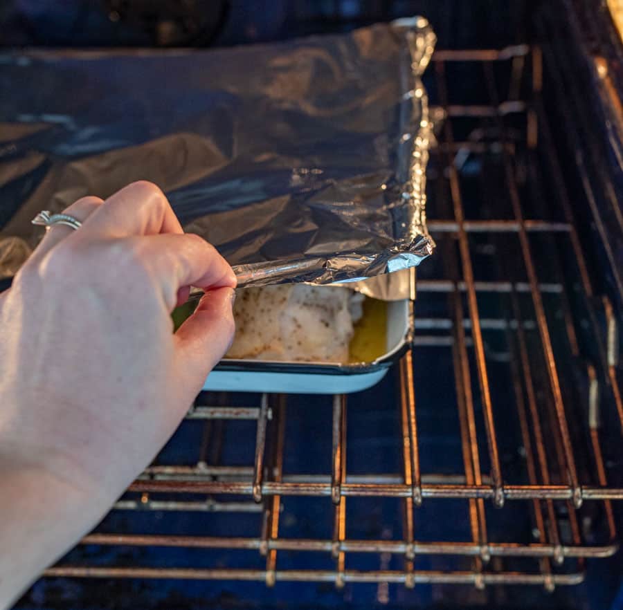 a hand checking the foiled baking dish that's inside an oven to slightly reveal the chicken thighs