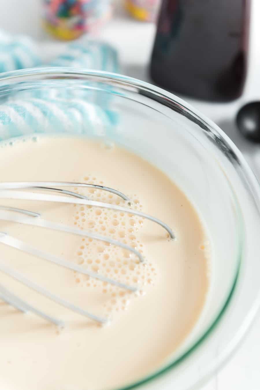 A metal whisk in a glass bowl of light tan batter, with bubbles forming on the surface—perhaps the start of a Snow Ice Cream Recipe. In the blurred background, a blue striped cloth and colorful objects add charm.