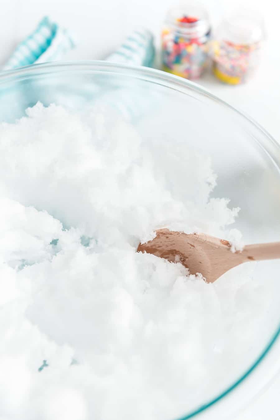 A clear glass bowl filled with fluffy white snow is being stirred with a wooden spoon, creating a delicious Snow Ice Cream Recipe. In the background, two jars of colorful sprinkles and a blue-striped cloth add a playful touch.