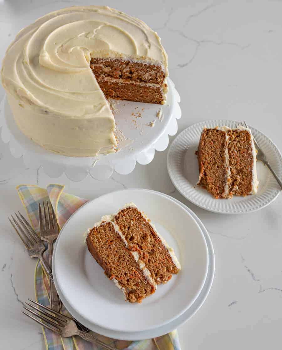 carrot cake with slice missing sitting on white cake stand with two slices of cake on round white plates on white surface with forks
