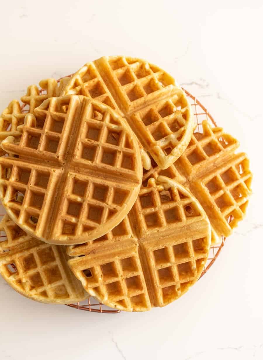 A stack of golden, round waffles made from a sourdough waffle recipe sits on a wire cooling rack against a white marble background.