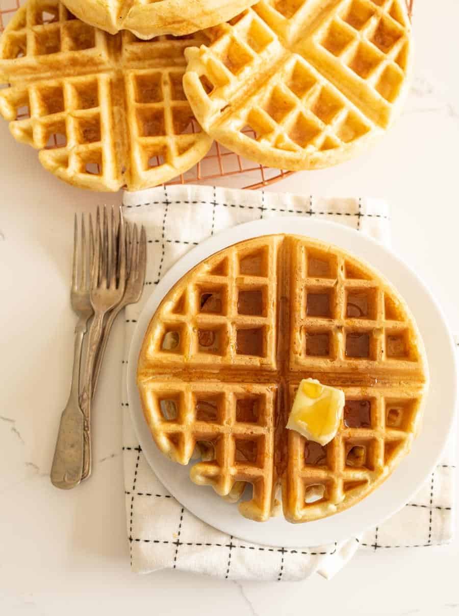 sourdough waffle on round white plate with pat of butter on white towel with forks.