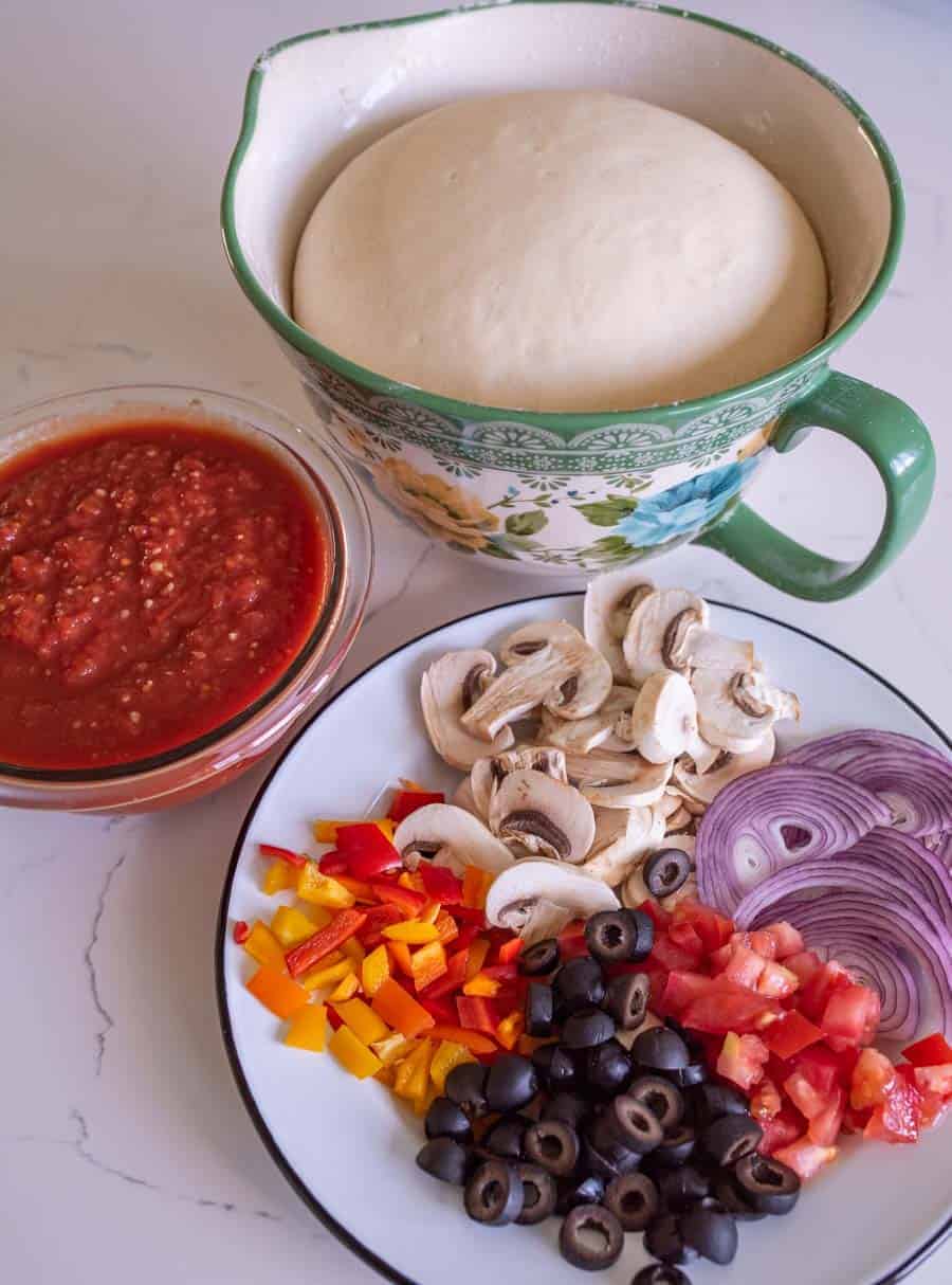 plate of olives tomato red onion bell pepper and mushrooms with pizza sauce and dough rising.