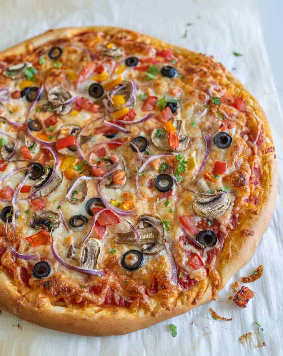 A baked veggie pizza recipe topped with melted cheese, sliced black olives, red onion, mushrooms, diced tomatoes, and chopped herbs, resting on a sheet of parchment paper.
